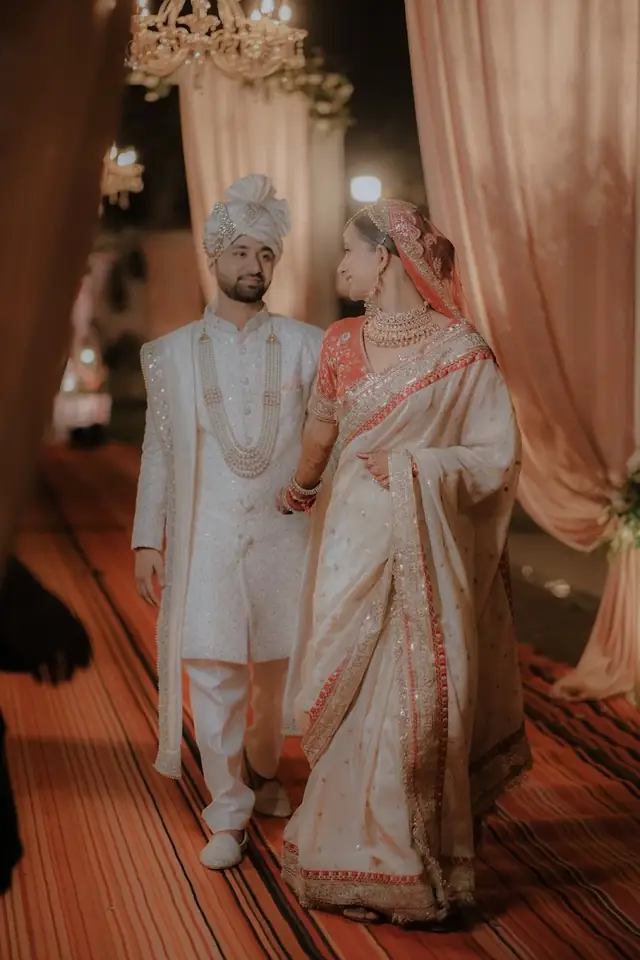 A couple in ivory walking down a draped aisle together, the bride holding her dupatta, floral arch overhead.