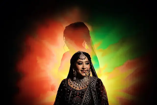 A bride in a dark embroidered lehenga and heavy kundan jewellery lit from behind with orange and green light and haze.
