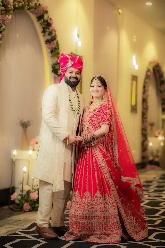 A groom in a pink turban and cream sherwani holding hands with the bride in a red lehenga, flower-covered arches down the hall behind them.