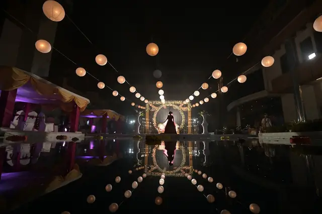 A wide night frame of a venue: strings of warm globe lights over still water, their reflection doubling the scene, a couple small at the centre.